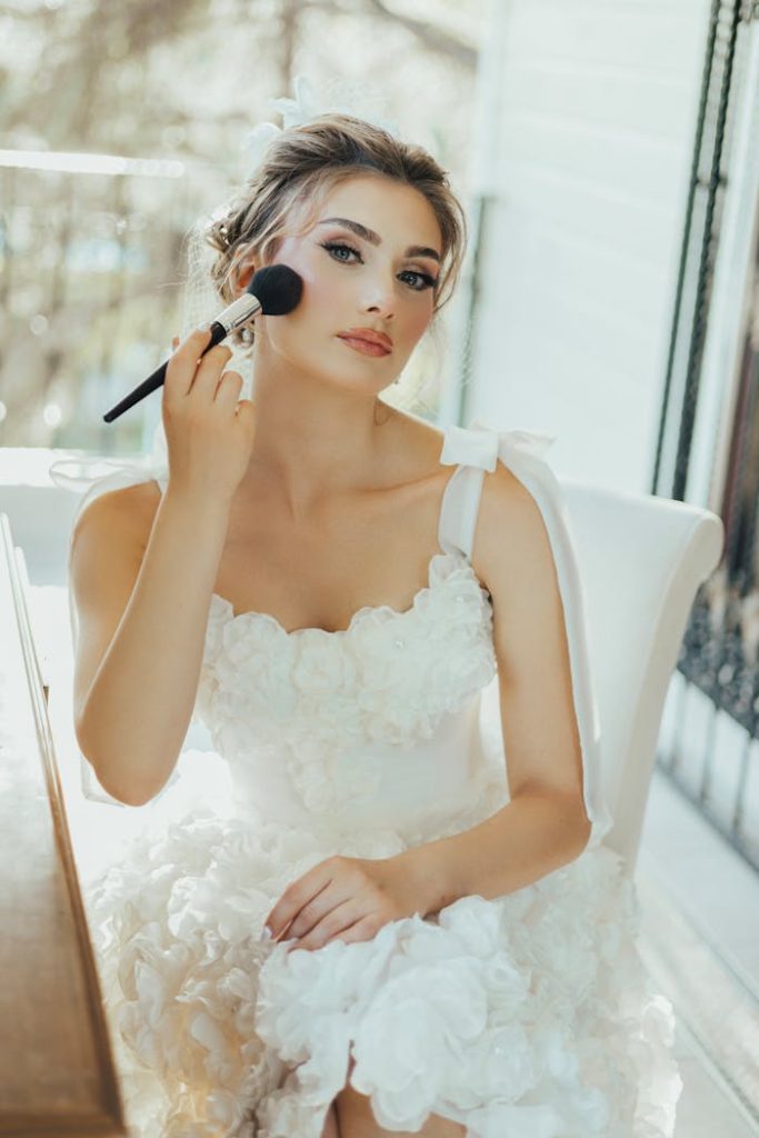 A young woman in a white wedding dress applying makeup with a brush indoors.